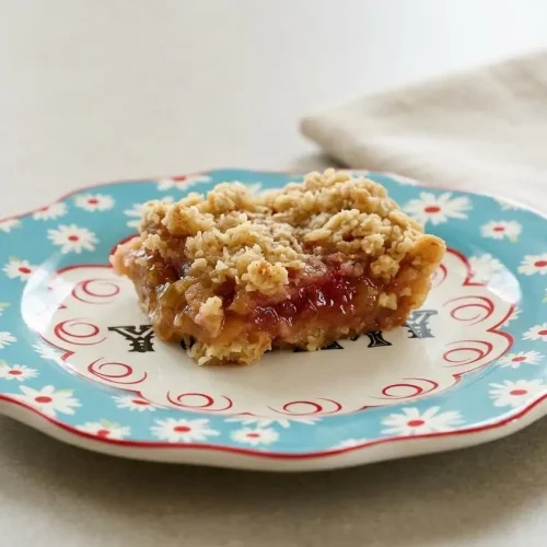Rhubarb bars recipe shown sliced in a glass baking dish, with a thick pink filling and a golden buttery oat crumble top.