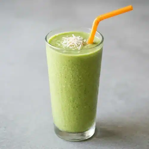 A finished Skinny Green Tropical Smoothie in a tall glass, showing a creamy pale green texture, surrounded by fresh pineapple, spinach, and