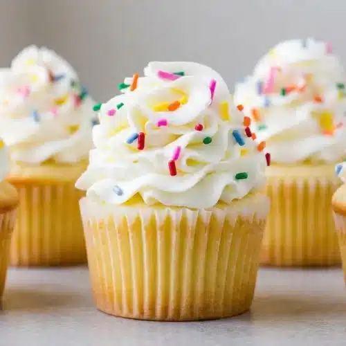 A perfect vanilla cupcake with a lightly golden top, resting in a simple paper liner on a dark surface.