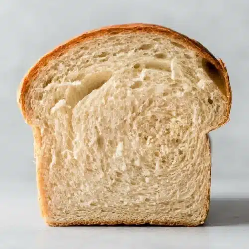 A golden-brown loaf from a white bread recipe, with one thick slice cut to show the soft, fluffy interior crumb, resting on a wooden board.