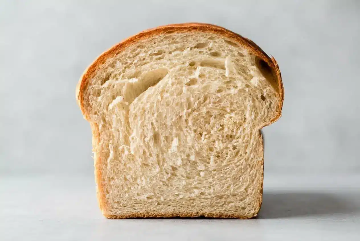 A golden-brown loaf from a white bread recipe, with one thick slice cut to show the soft, fluffy interior crumb, resting on a wooden board.