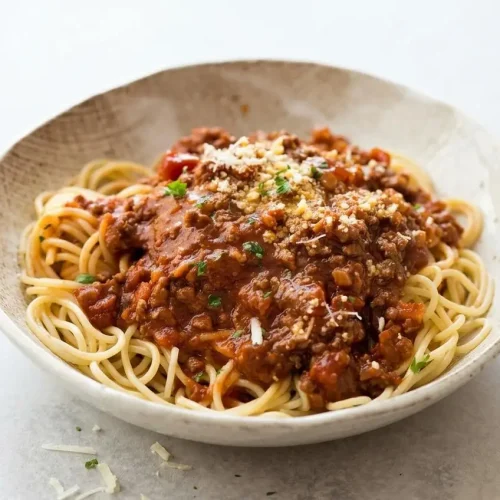 A white bowl of Spaghetti Bolognese with a rich, glossy meat sauce clinging to the pasta, finished with fresh parsley and Parmesan cheese.