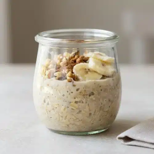 A glass jar of Tres Leches Overnight Oats, topped with fresh banana slices, chopped walnuts, and a dusting of cinnamon.