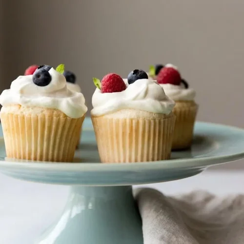 Frosted Angel Food Cupcakes topped with pristine whipped cream and fresh berries in a clean kitchen setting.