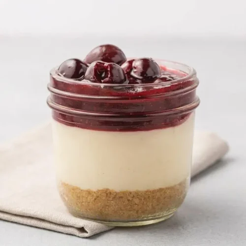 Individual Cherry Cheesecake In a jar showing a buttery graham crust, smooth cream cheese filling, and glossy cherry topping.