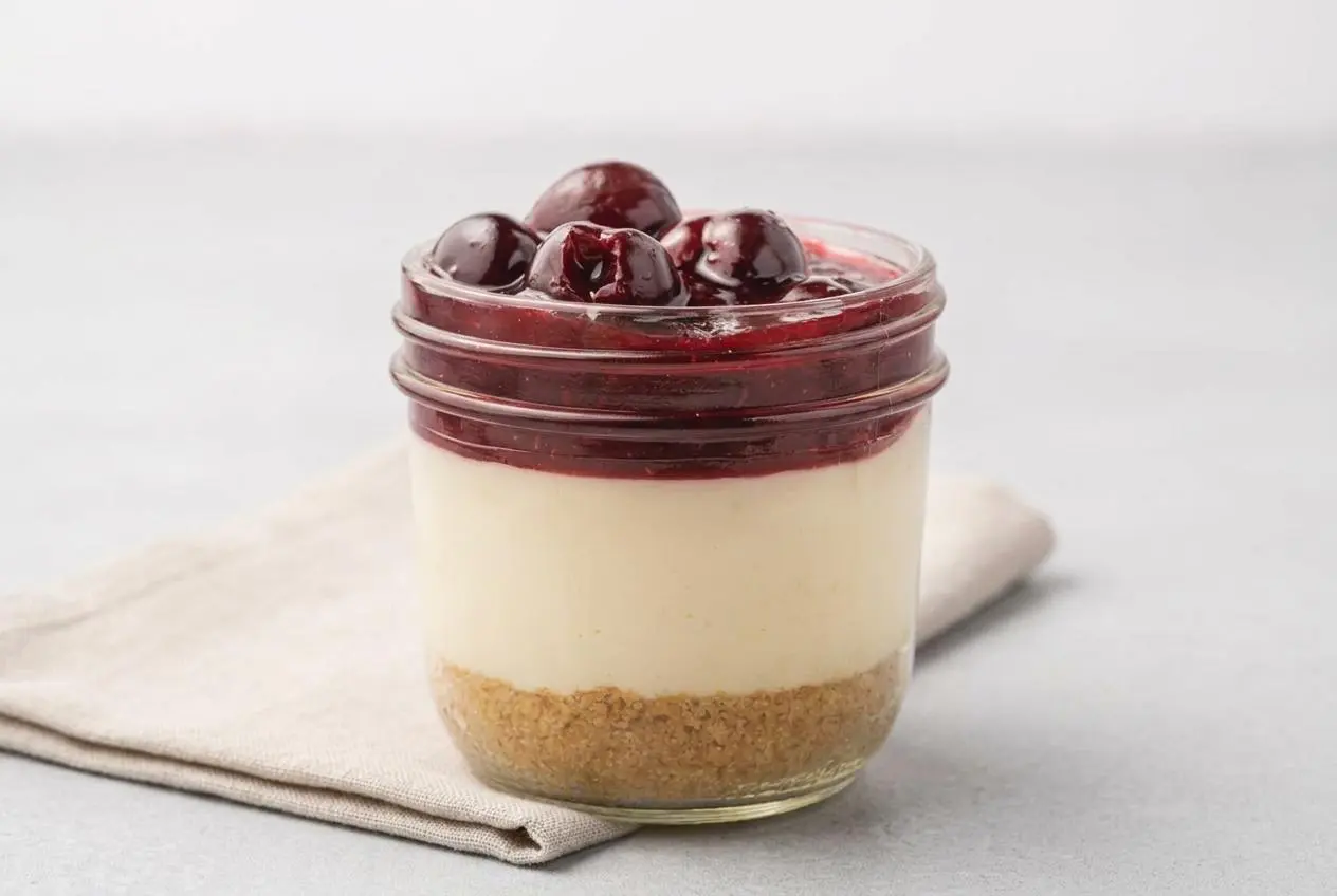 Individual Cherry Cheesecake In a jar showing a buttery graham crust, smooth cream cheese filling, and glossy cherry topping.