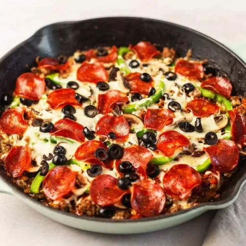 Baked crustless pizza in a skillet showing bubbly melted mozzarella, turkey pepperoni, black olives, and golden-brown edges.