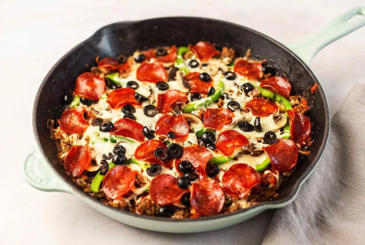 Baked crustless pizza in a skillet showing bubbly melted mozzarella, turkey pepperoni, black olives, and golden-brown edges.
