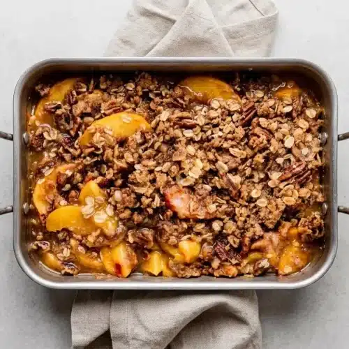 Baked peach crisp recipe with a golden brown oat pecan topping in a deep dish ceramic baking pan.