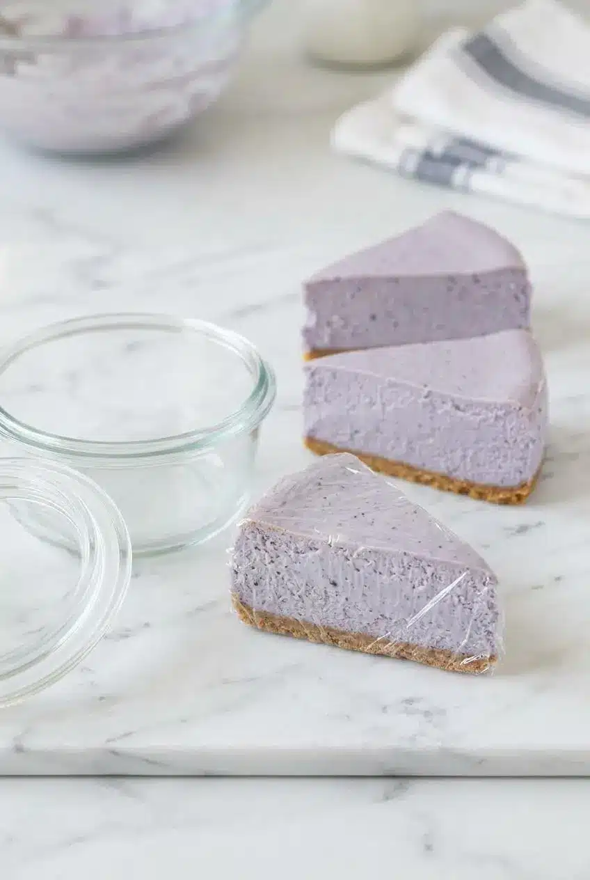 A slice of purple lavender cheesecake wrapped tightly in plastic wrap next to an airtight glass container.