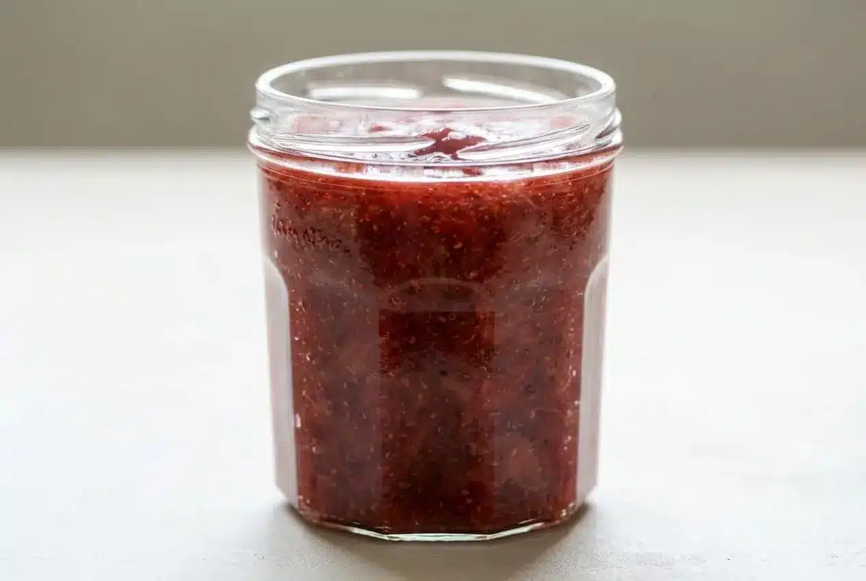 A small glass jar filled with glossy strawberry rhubarb chia jam showing visible fruit pieces and tiny chia seeds.