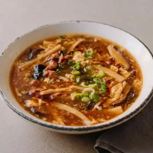 A bowl of hot and sour soup showing glossy mahogany broth with egg ribbons, firm tofu, and wood ear mushrooms.