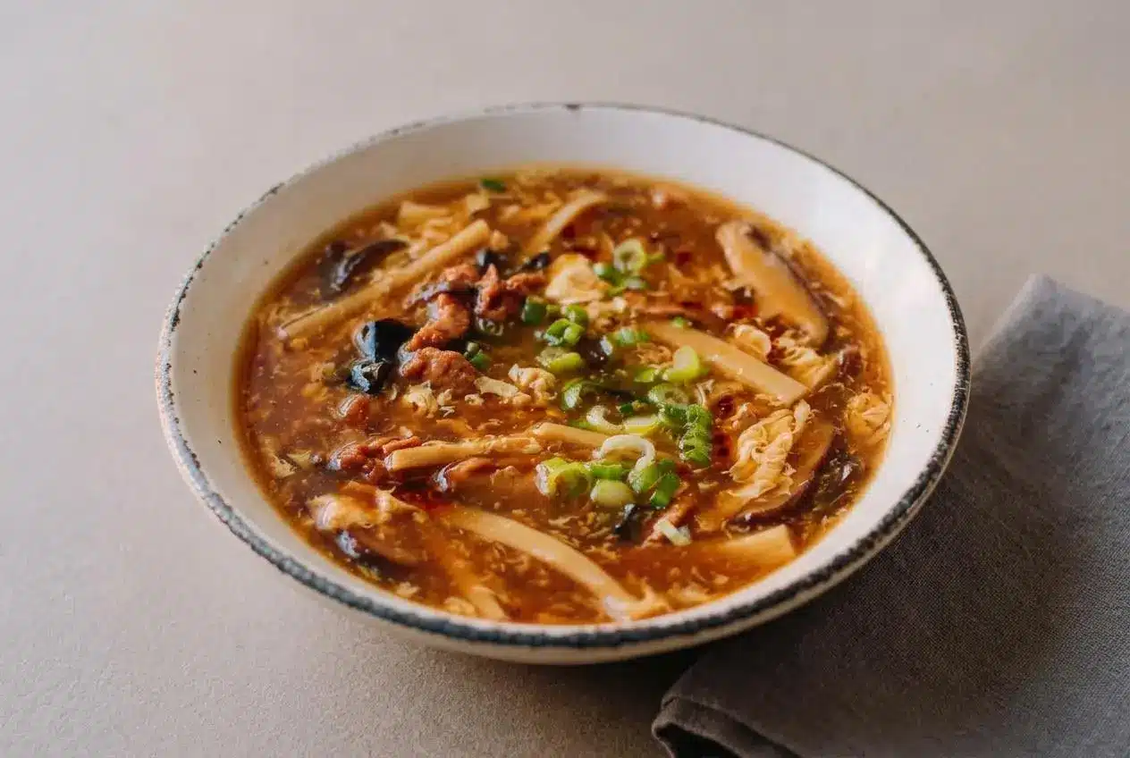 A bowl of hot and sour soup showing glossy mahogany broth with egg ribbons, firm tofu, and wood ear mushrooms.