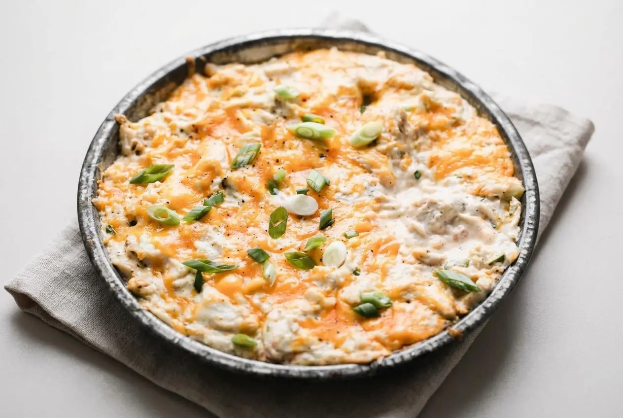 Baked crab dip recipe in a round dish with a golden bubbly cheddar crust and fresh green onions.