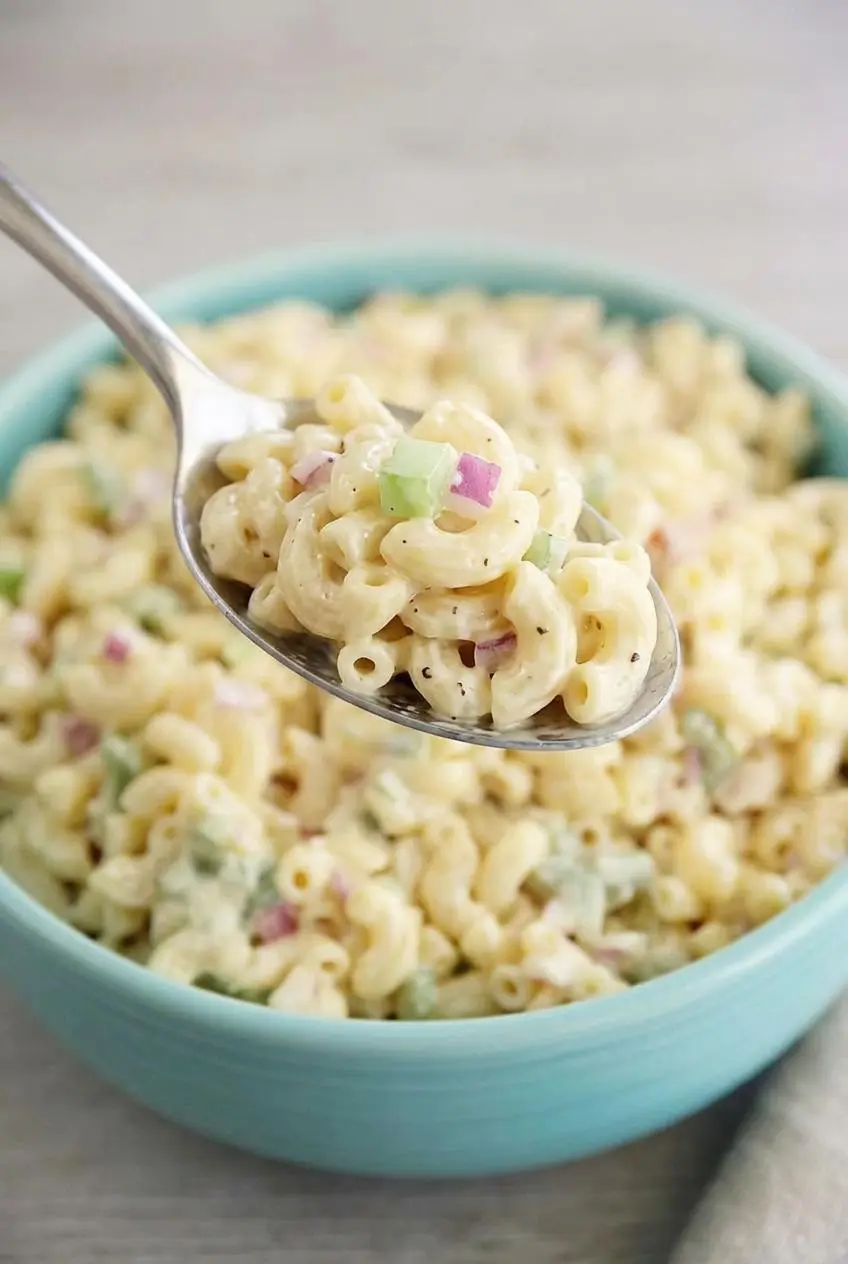 A spoonful of glossy macaroni salad lifted over a bowl, showing the creamy mayonnaise dressing coating the pasta.