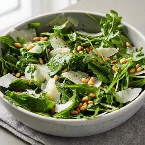 Freshly tossed arugula salad with shaved Parmesan cheese and toasted pine nuts in a rustic ceramic bowl.