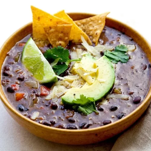 Hearty black bean soup in a ceramic bowl garnished with fresh cilantro, diced avocado, and a lime wedge.