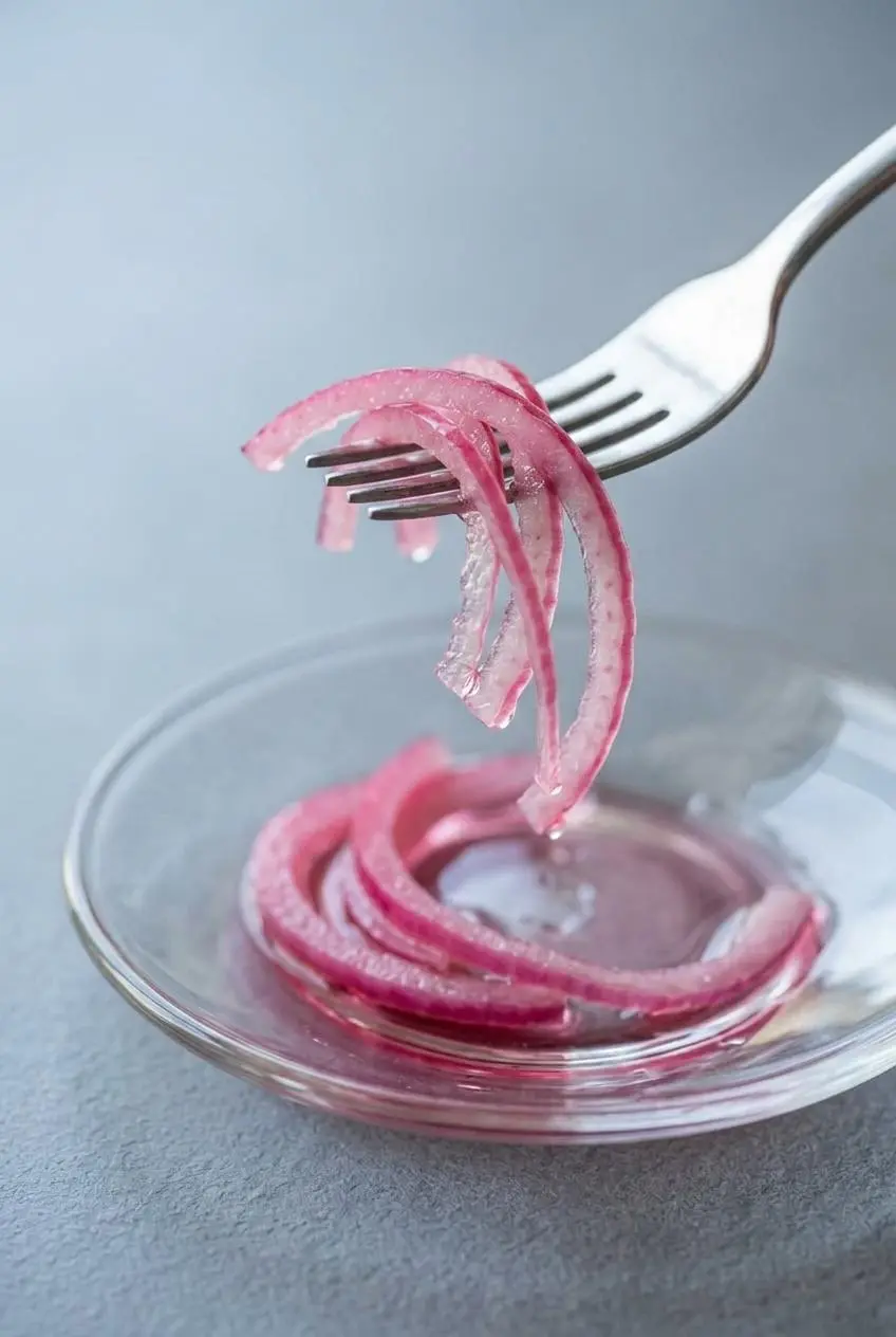 Paper-thin, translucent pink pickled red onion strands lifted on a silver fork over a small glass saucer.