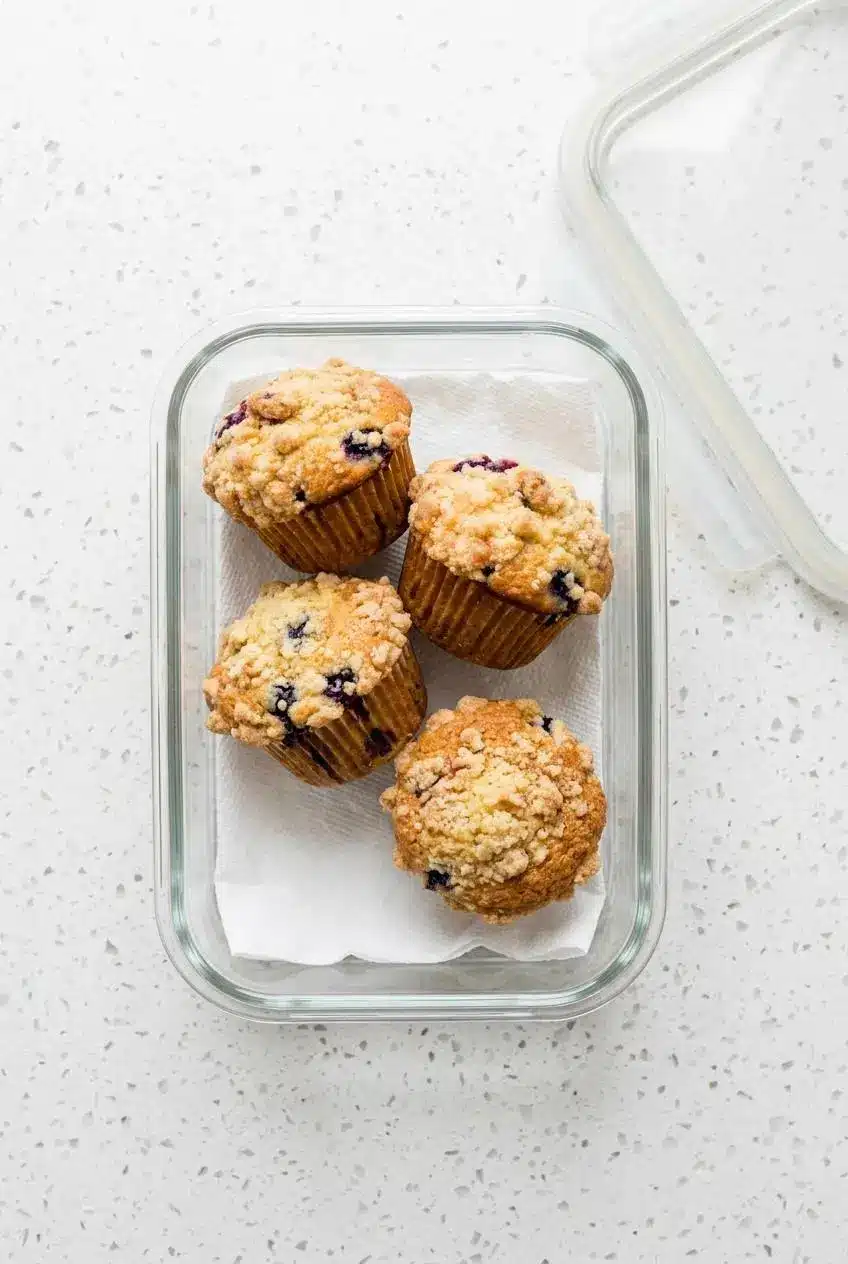 Four cooled muffins stored in a glass airtight container lined with paper towels to absorb moisture.