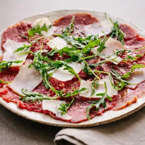 Plated beef carpaccio with paper-thin raw tenderloin, shaved Parmigiano, peppery arugula, and a glossy olive oil drizzle.