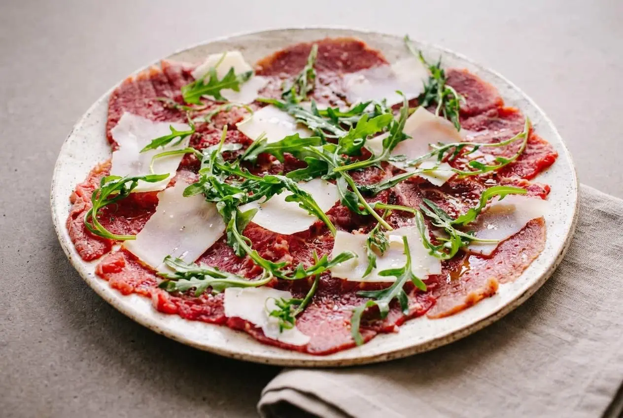 Plated beef carpaccio with paper-thin raw tenderloin, shaved Parmigiano, peppery arugula, and a glossy olive oil drizzle.