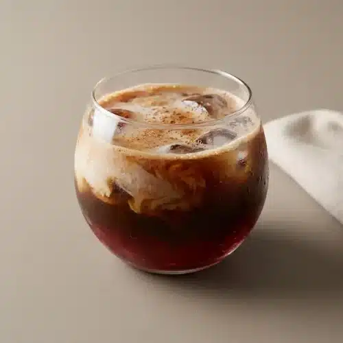 Iced cranberry coffee poured over ice with a light frothy foam layer and a soft rosy amber hue.