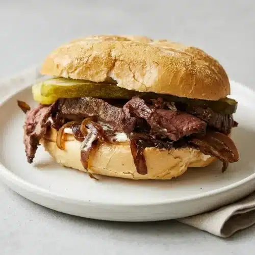 Juicy roast beef sandwich layered with tender sliced beef, caramelized onions, and pickles on a toasted Kaiser roll.