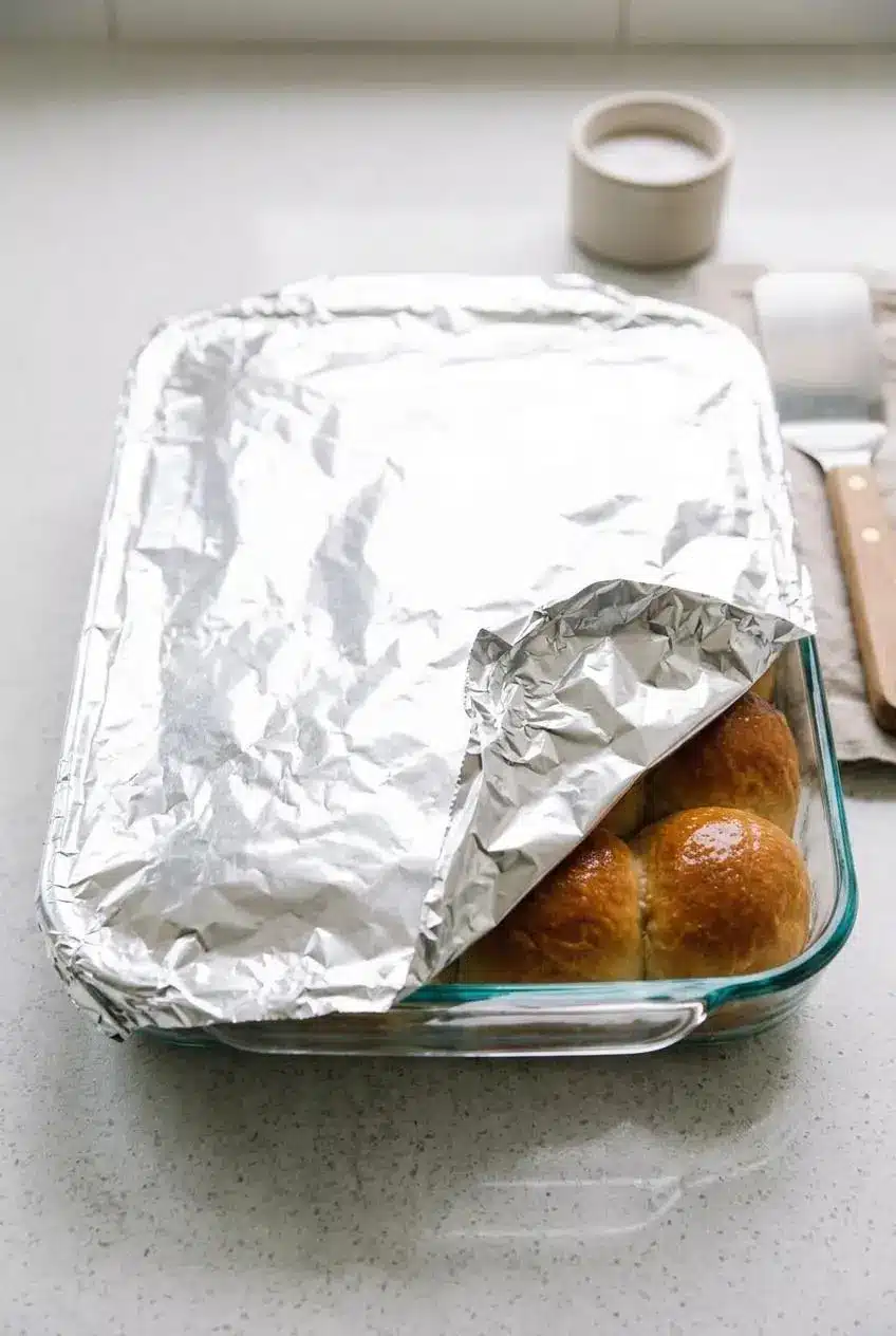 Pan of baked dinner rolls loosely covered with foil on a kitchen counter.
