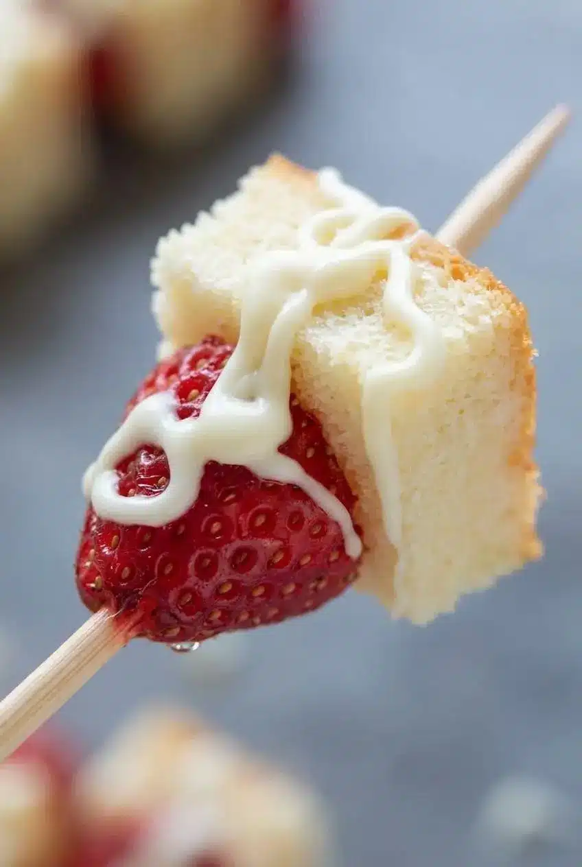 Close-up of white chocolate drizzle set over a fresh strawberry and tender vanilla cake cube on a wooden skewer.