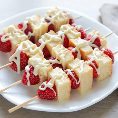 Strawberry shortcake kabobs with golden cake cubes, fresh berries, and a set white chocolate drizzle on a platter.
