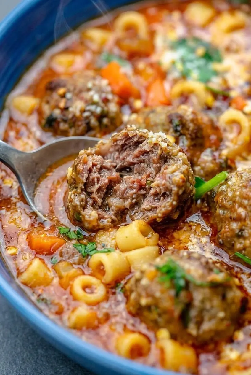 Spoon breaking open a tender, juicy mini meatball inside a bowl of tomato broth with ditalini pasta.