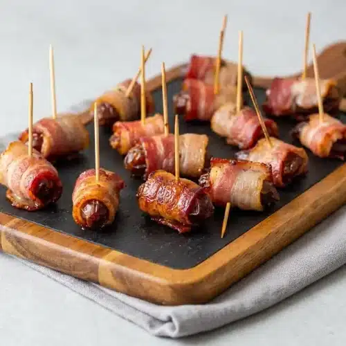 Bacon Wrapped Dates baked crisp with caramelized edges and secured with wooden toothpicks on a platter.