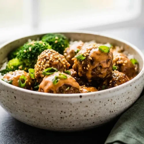 Baked firecracker meatballs with golden edges, creamy sriracha sauce drizzle, sliced green onions, and sesame seeds.