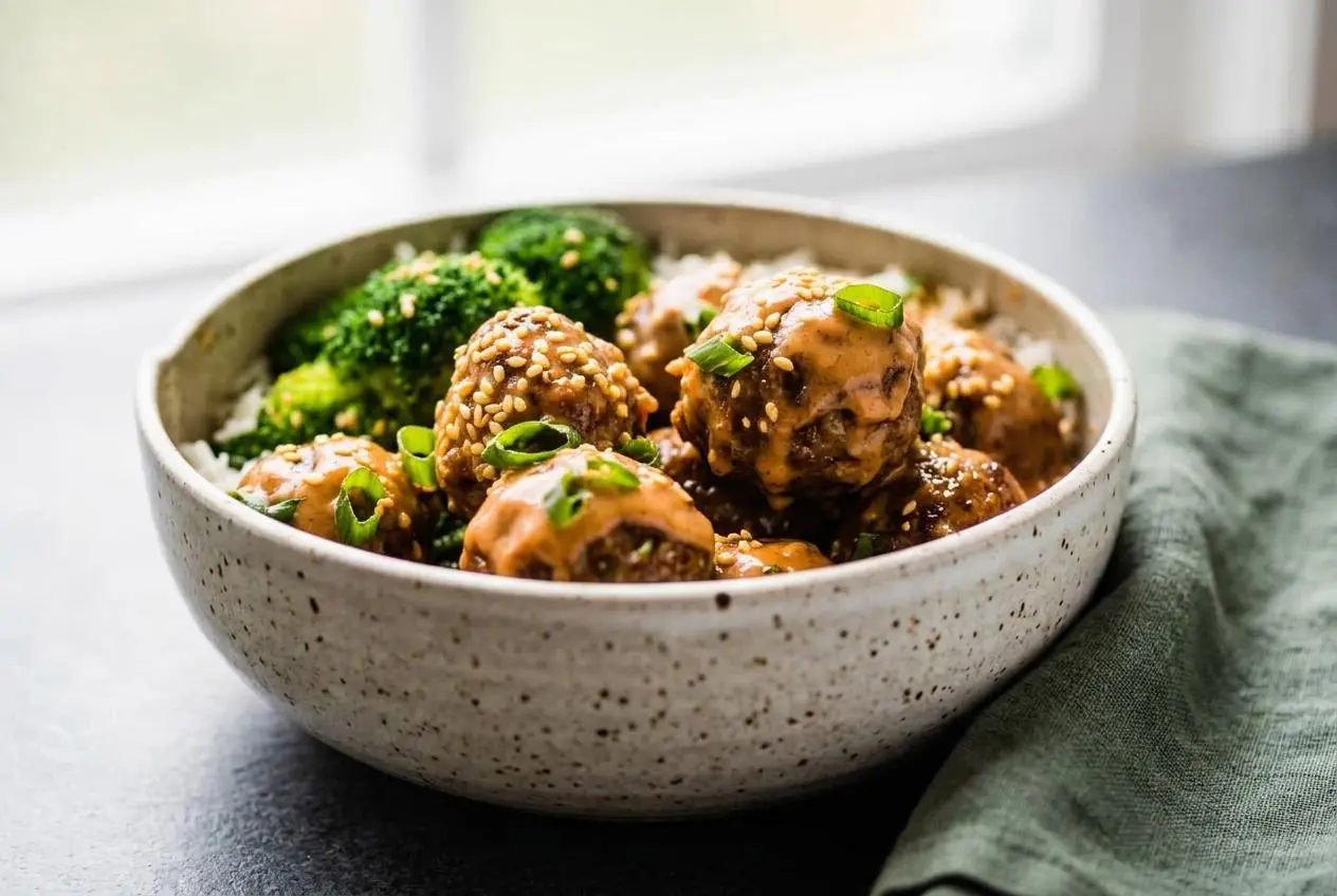 Baked firecracker meatballs with golden edges, creamy sriracha sauce drizzle, sliced green onions, and sesame seeds.