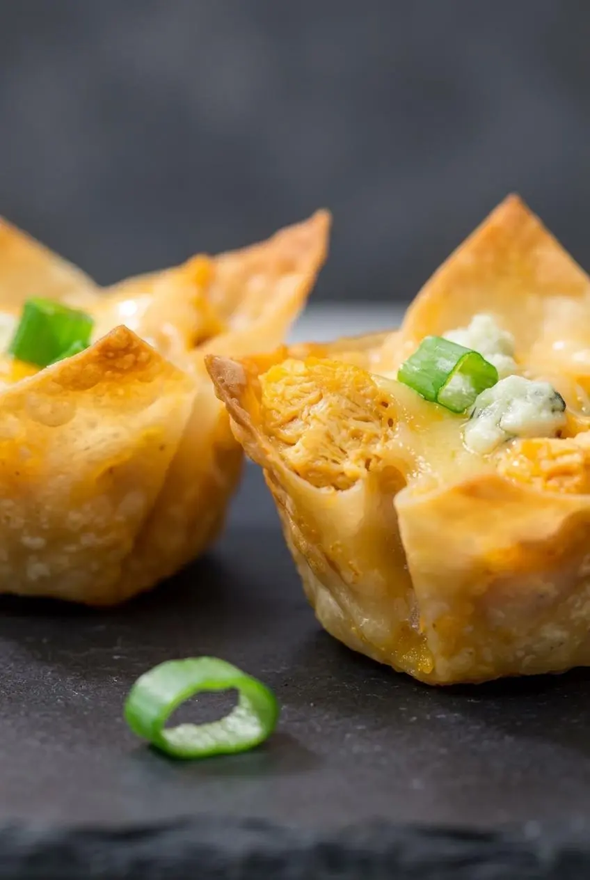 Close-up of two buffalo chicken wonton cups on dark slate showing crisp golden corners and melted cheese.