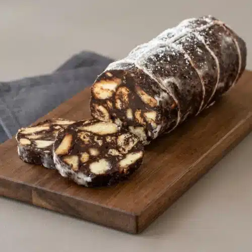 Chocolate salami dusted with powdered sugar and sliced into marbled rounds showing biscuit pieces.