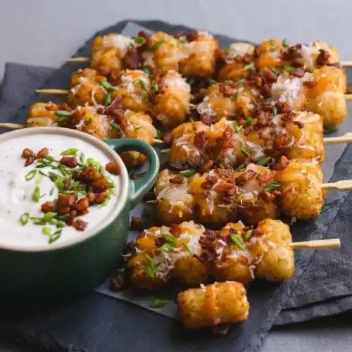 Crispy loaded tater tot skewers topped with melted cheese, chopped bacon, and green onions on a platter with ranch dip