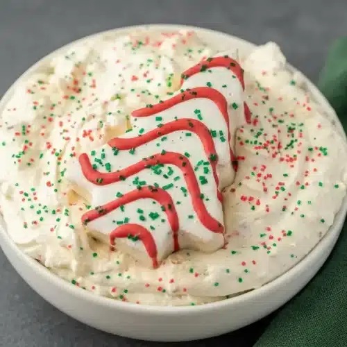 Fluffy white christmas tree cake dip in a bowl topped with red and green sprinkles and a whole snack cake.