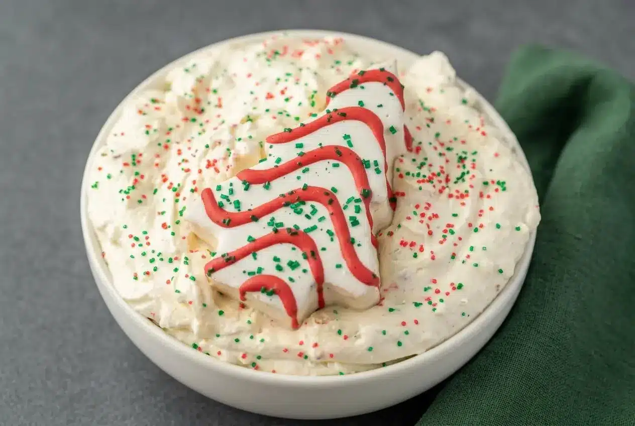 Fluffy white christmas tree cake dip in a bowl topped with red and green sprinkles and a whole snack cake.
