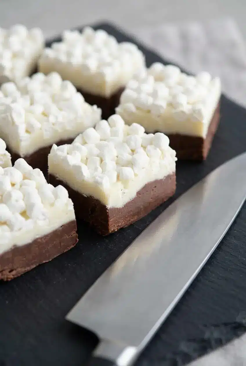 Close-up of sliced chocolate fudge squares on dark slate next to a chef knife showing clean dark and white layers