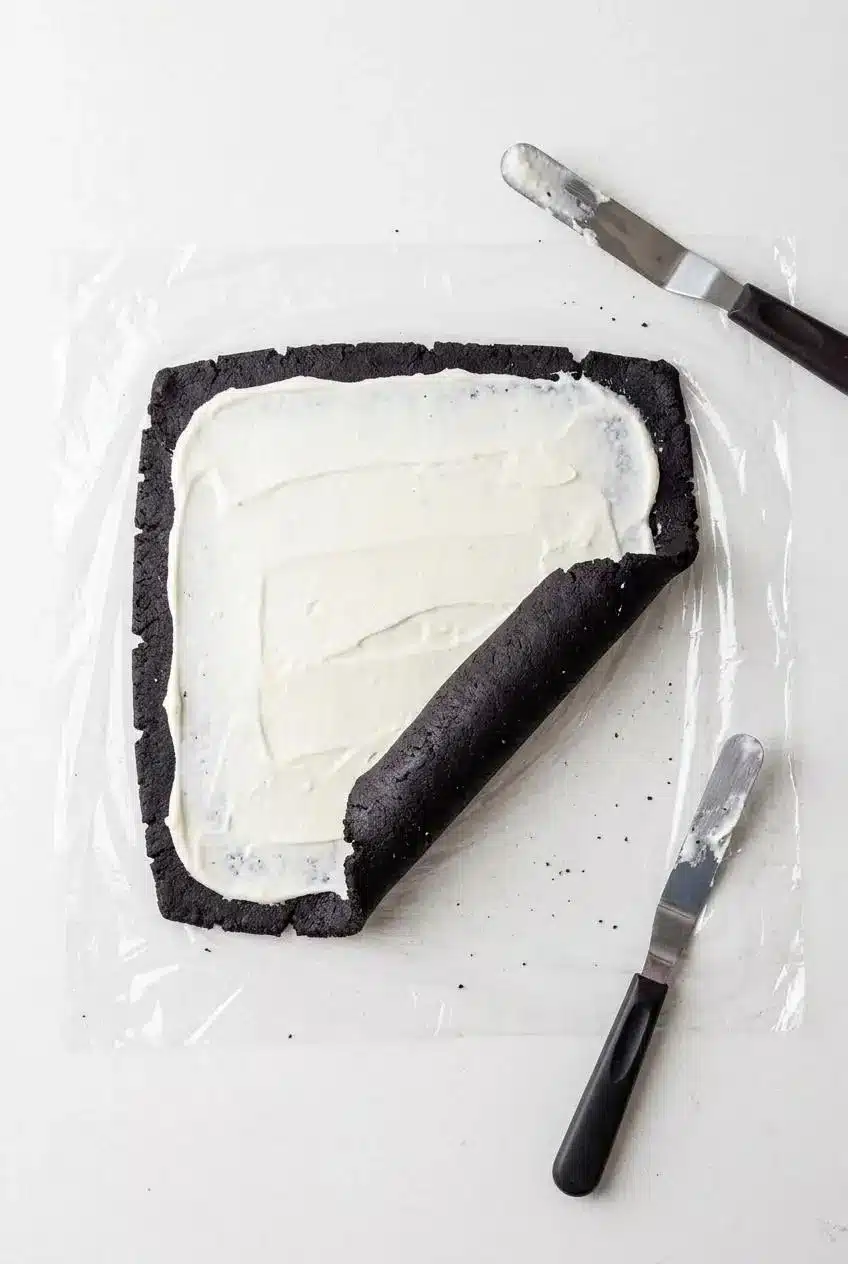 Overhead view of chocolate cookie dough rolled on plastic wrap, spread with white cream filling, and curling into a log.