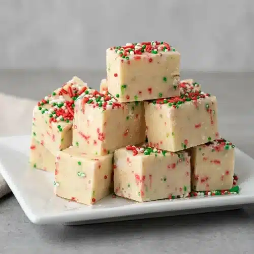 Square slices of creamy sugar cookie fudge topped with festive holiday sprinkles on a dessert plate.